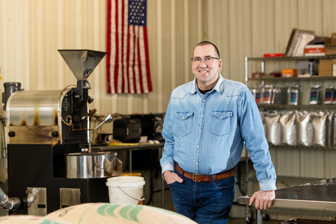 Man standing in a coffee shop with coffee equipment and an American flag in the background